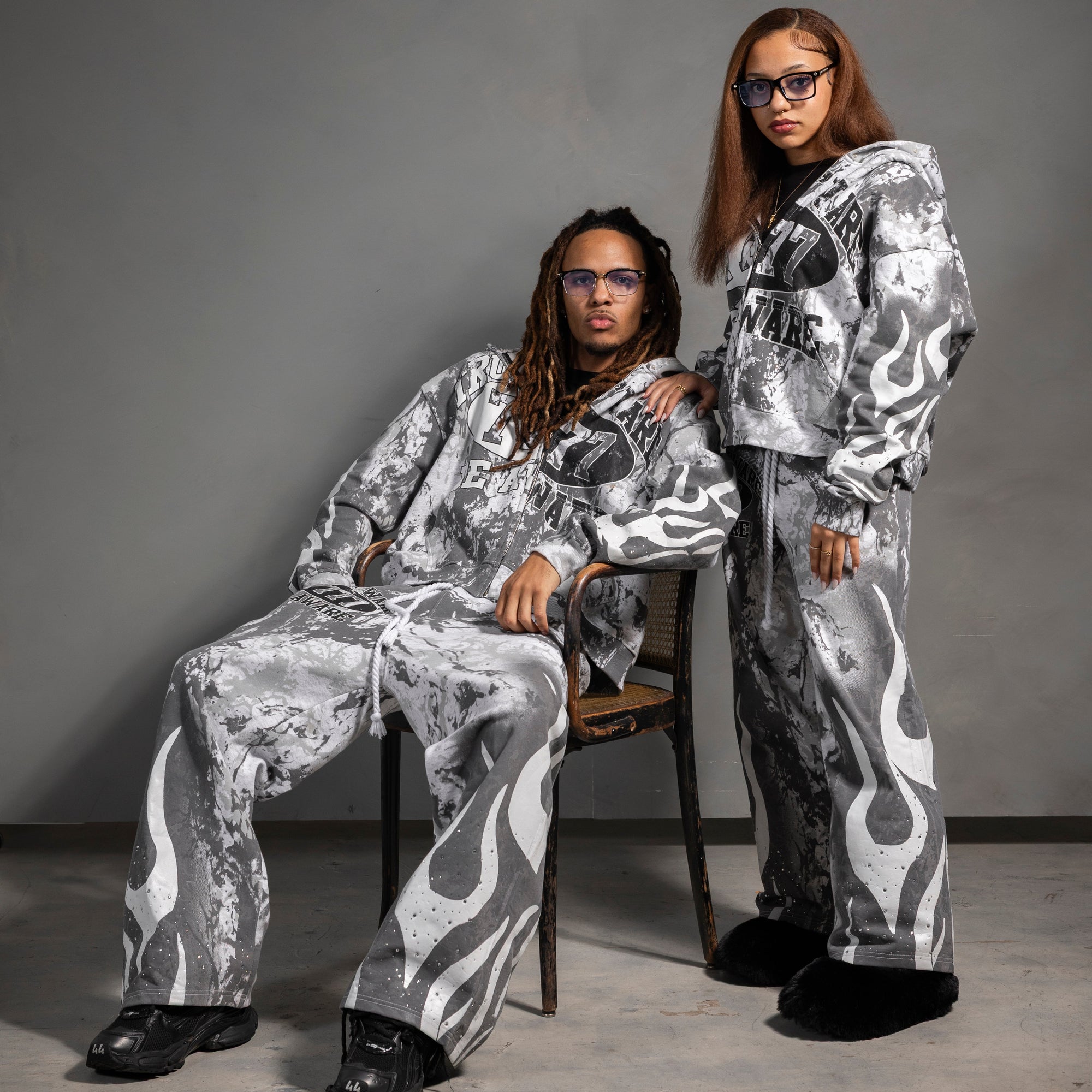 Two people wearing matching silver and gray patterned outfits against a gray background.